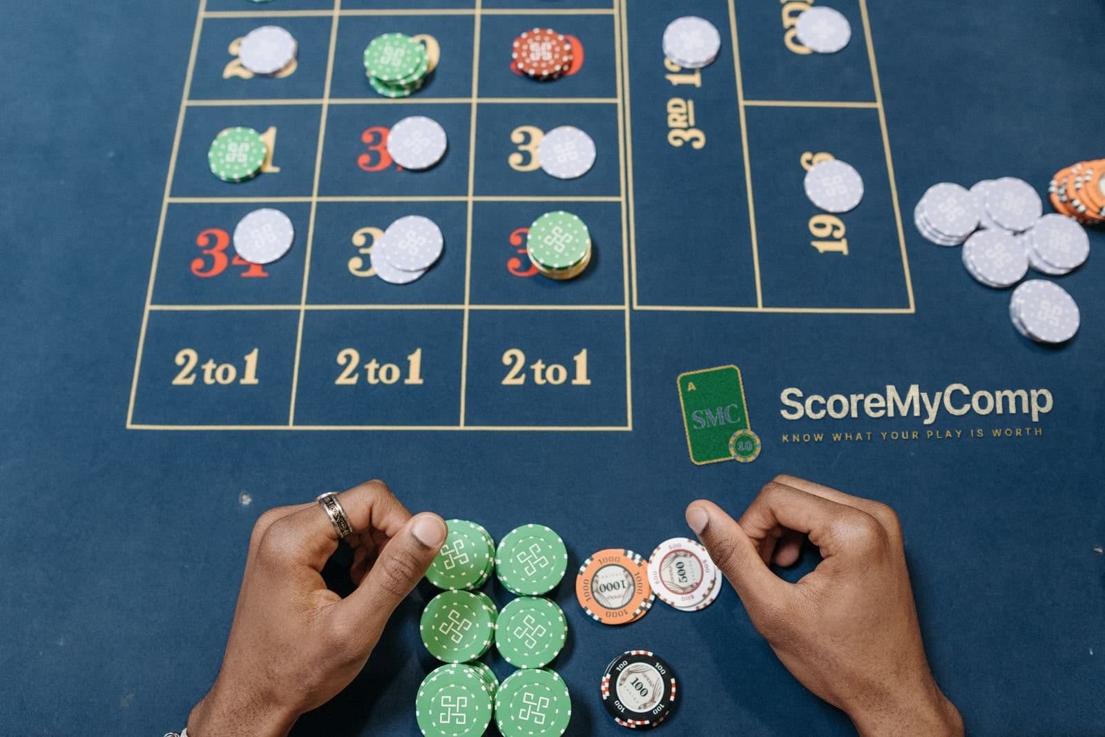 Players placing chips on a casino table layout with the ScoreMyComp logo printed on the felt