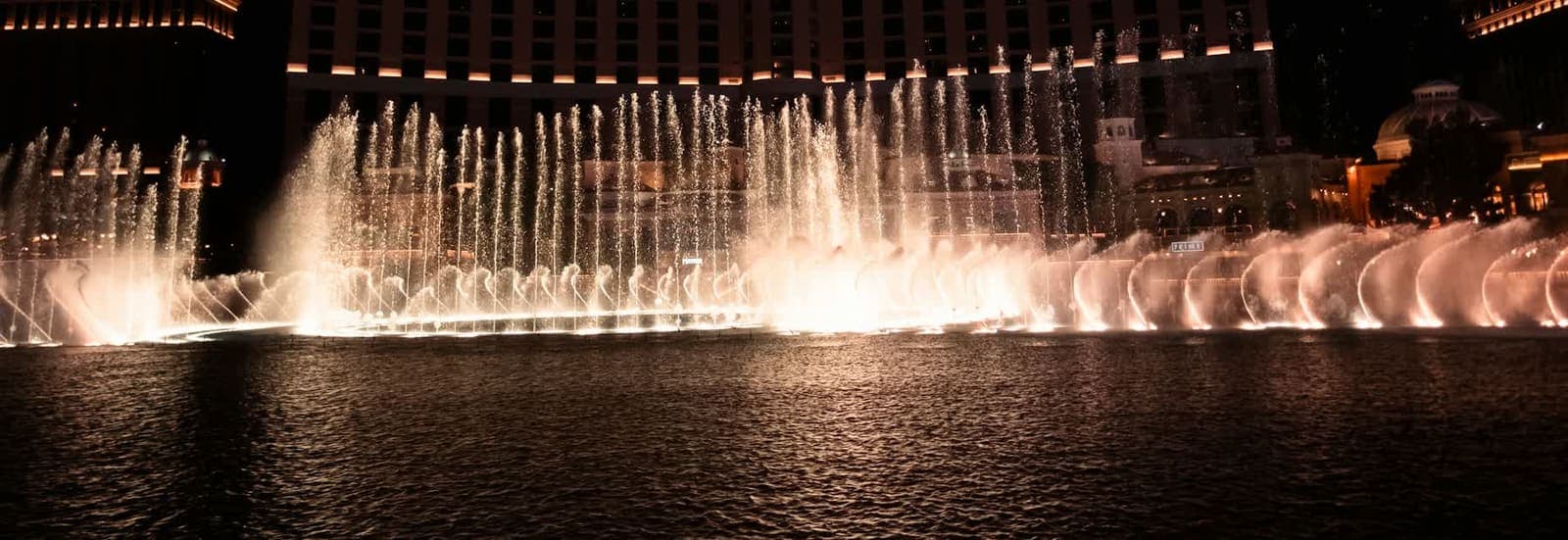 Fountain jets rising from a lake at night, with the hotel name cropped out of the frame.