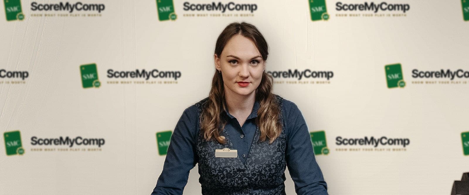 Blackjack dealer at a casino table in front of a ScoreMyComp logo backdrop
